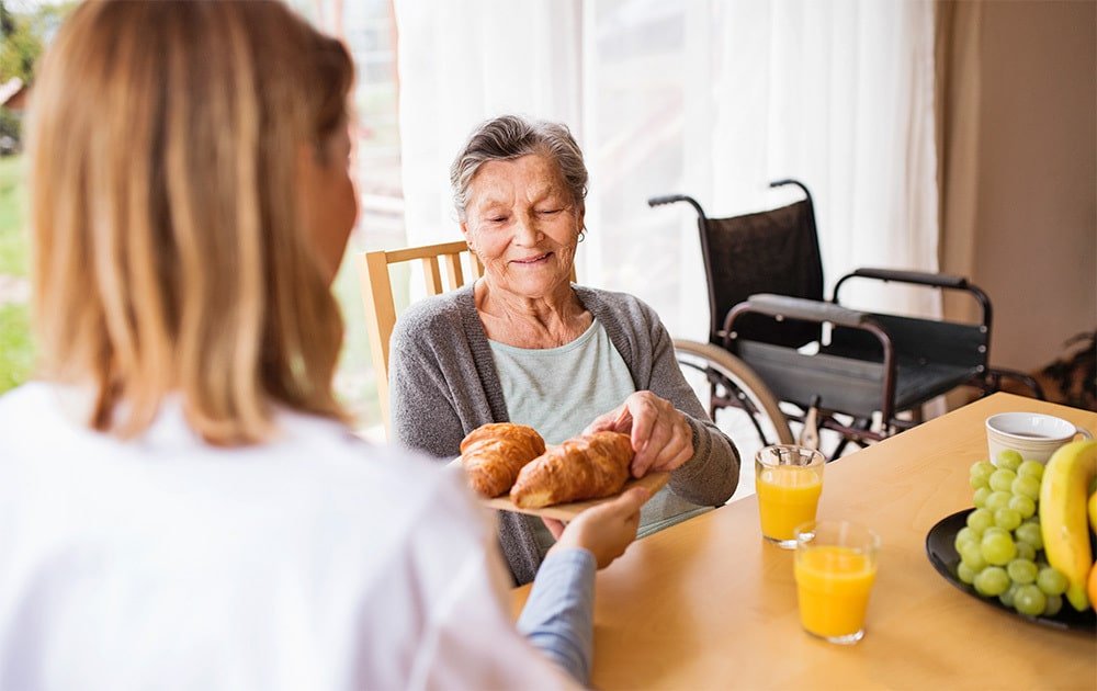 Health visitor and a senior woman during home visit. A nurse giving food to an elderly woman sitting at the table.